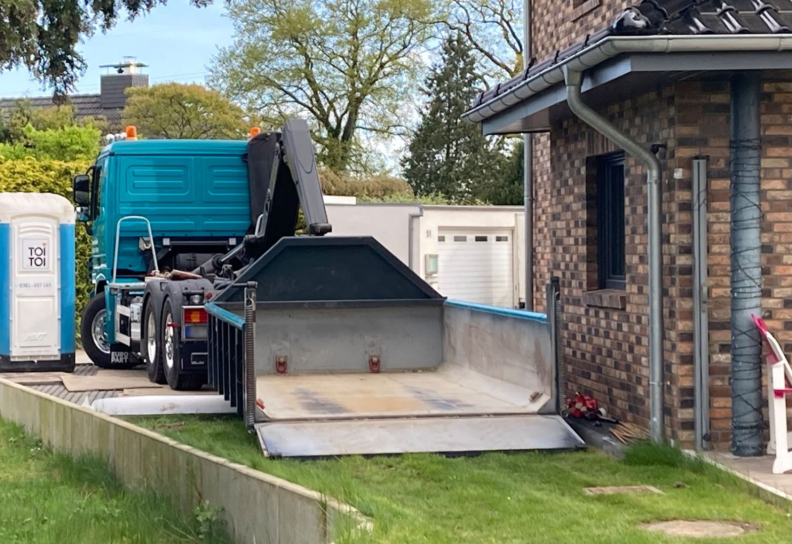 Türkiser LKW des Containerdienstes Käselau setzt einen Abrollcontainer mit geöffneter Rampe direkt vor einem Wohnhaus ab