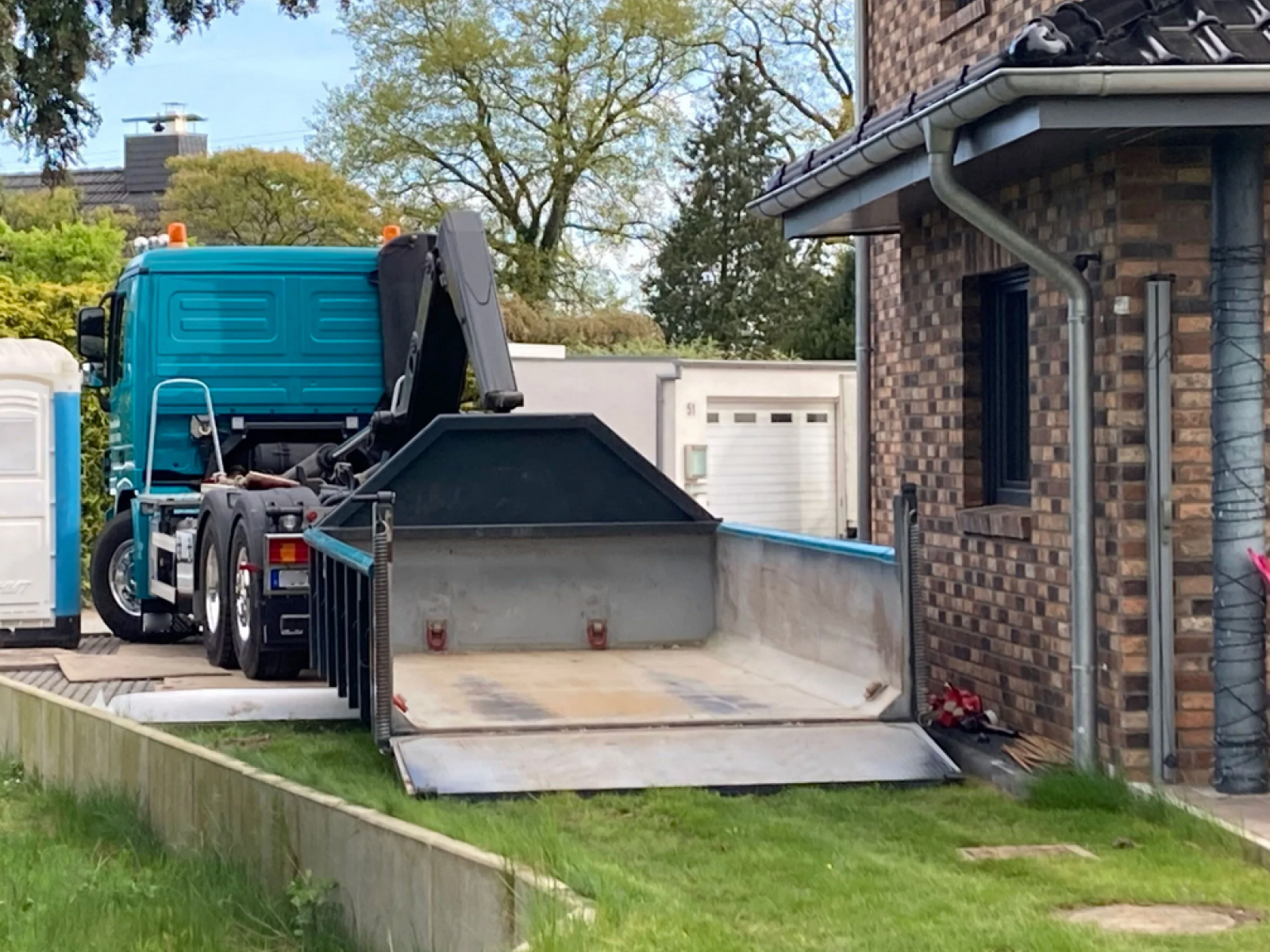 Türkiser LKW des Containerdienstes Käselau setzt einen Abrollcontainer mit geöffneter Rampe direkt vor einem Wohnhaus ab