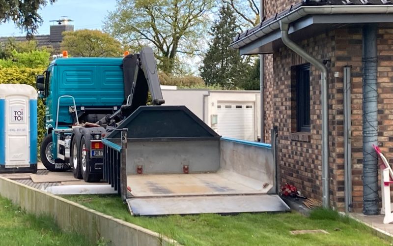 Türkiser LKW des Containerdienstes Käselau setzt einen Abrollcontainer mit geöffneter Rampe direkt vor einem Wohnhaus ab